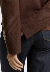 Bruine ribgebreide trui met een hoge hals, voorzien van lange mouwen en een relaxed fit. Gepaard met donkere denim jeans.