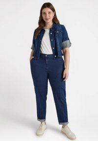 Jasnoblue kurtka dżinsowa o skróconym kroju i podwiniętych rękawach, zestawiona z ciemnoniebieskimi spodniami o wysokim stanie. Modelka w białej bluzce i beżowych sneakerach.