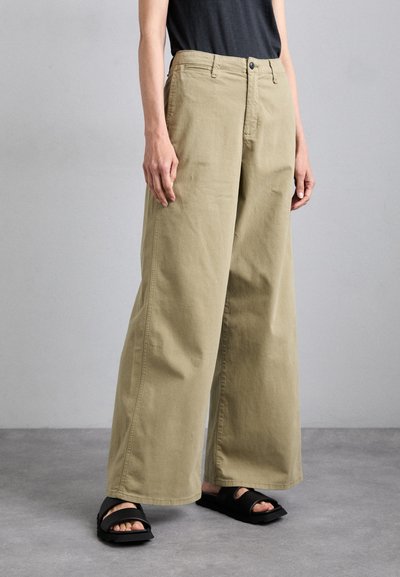 Pantalon large en mélange de coton couleur kaki, avec une taille à boutons et des poches latérales, associé à des sandales noires à enfiler.