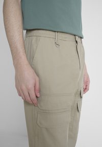 Beige cargobroeken met meerdere zakdetails, een elastische tailleband en een zijclip. De stof heeft een gladde textuur en een lichte uitstraling.