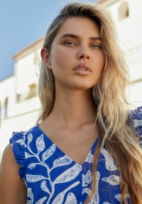 Mladá žena s dlouhými blond vlasy, oblečená v modrých šatech s bílými vzory listů, stojící venku poblíž bílých budov ve středomořském stylu.