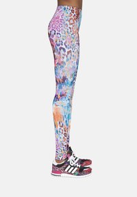 Kolorowe legginsy z jaskrawym wzorem w panterkę w odcieniach fioletu, niebieskiego i pomarańczowego, zestawione z wielokolorowymi butami sportowymi z czarnymi akcentami.
