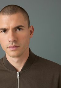 Chaqueta bomber marrón con cierre de cremallera, cuello acanalado y tejido texturizado. El modelo tiene el cabello corto, con un fondo de tono neutro.