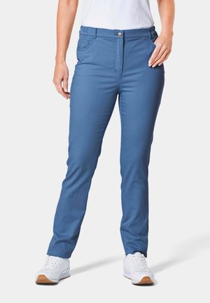Pantalons en tissu extensible bleu, avec poches avant, une fermeture éclair et un bouton. Conception à jambes effilées, portés avec des chaussures de sport blanches.
