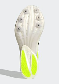 Valkoisen urheilukengän pohja, jossa mustia yksityiskohtia, kuusi hopeista nastaa ja teksturoitu vihreä pitoalue. Siinä on virtaviivainen muotoilu ja joustavat urat.