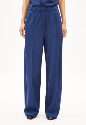 Pantaloni blue a gamba larga con una texture liscia e una vita elasticizzata. Presentano pince frontali e tasche laterali, abbinati a scarpe nere.