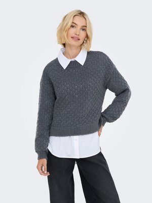 Donna bionda che indossa un maglione grigio a maglia strutturata sopra una camicia bianca con colletto, abbinato a pantaloni neri larghi, che posa con una mano sul fianco.