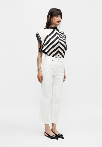 Γυναίκα φοράει πουκάμισο με σχέδιο σχήματος ζιγκ-ζαγκ σε ασπρόμαυρο, λευκό παντελόνι με ψηλή μέση και μαύρα μυτερά παπούτσια slingback με ασημένια πόρπη.