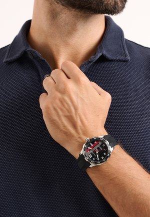 Férfi fekete texturált galléros pólóban, bal kézzel igazítja a gallérját, bal csuklóján fekete-ezüst karórát visel.