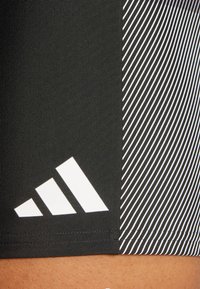 Czarne krótkie spodenki sportowe z białym logo Adidas i teksturowaną tkaniną. Wzór zawiera ukośne białe paski po jednej stronie.