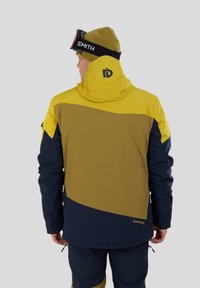 Giacca da neve gialla, marrone e blu navy. Presenta un cappuccio, un design bicolore e un logo sul retro. Realizzata in materiale resistente alle intemperie.