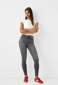 Szürke, skinny farmernadrág magas derékkal, egy formázott fehér pólóval és piros sportcipővel párosítva. Simító felület, enyhe kopási részletekkel.