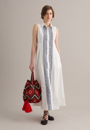 Vrouw draagt een mouwloze witte jurk met verticale zwarte borduursels, houdt een grote rode tas met patroon en kwastjes vast, staand tegen een effen achtergrond.