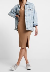 Kobieta w jasnoniebieskiej dżinsowej kurtce nosząca bezrękawową brązową sukienkę midi o prążkowanej fakturze z bocznym rozcięciem, w połączeniu z beżowymi trampkami.