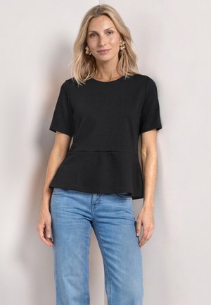 Vrouw met blond haar met gouden oorbellen, zwart kortmouw peplumtopje en lichtblauwe spijkerbroek, staand tegen een neutrale achtergrond.