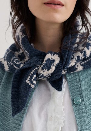 Gebreide sjaal in marineblauw en roomkleurig patroon om de nek gestrikt, gecombineerd met een lichtblauwe cardigan en een wit gerimpeld overhemd, met focus op textuur en detail.