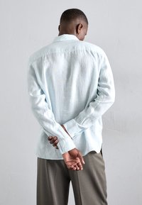 Camisa de lino azul claro con mangas largas, puños abotonados y un corte holgado, llevada con pantalones grises, vista desde atrás.