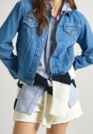 Person iført en blå denimjakke over en lyseblå skjorte med en sort-hvid strikket sweater bundet om livet og en cremefarvet nederdel.