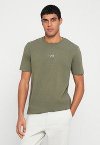 Oliwkowo-zielona koszulka z krótkim rękawem, okrągłym dekoltem i małym srebrnym logo na piersi, wykonana z gładkiej tkaniny.