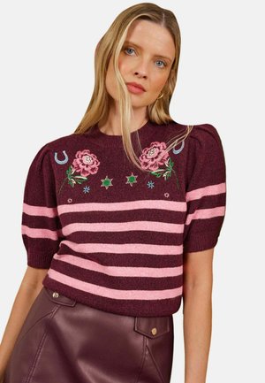 Kvinde med blondt hår iført en bordeaux og lyserød stribet sweater med blomstret broderi, kombineret med en mørk bordeaux lædernederdel.