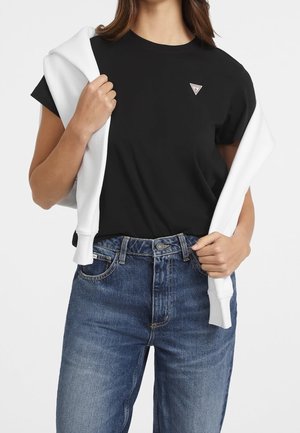 T-shirt nera a maniche corte con un piccolo logo triangolare, abbinata a jeans a vita alta di colore blu chiaro e un maglione bianco drappeggiato sulle spalle.