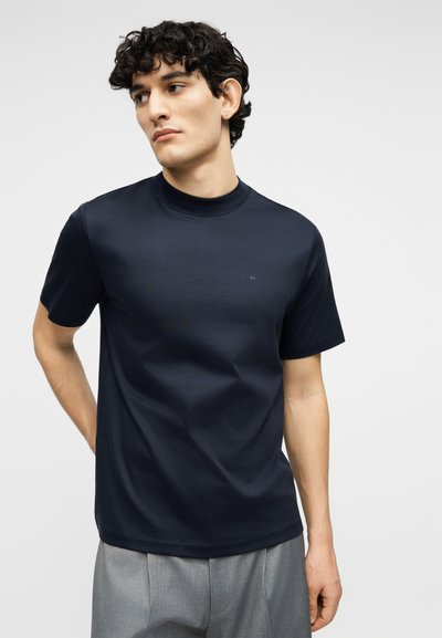 Νέος άνδρας με κοντά σγουρά μαλλιά, φορώντας μπλε μπλουζάκι με κοντά μανίκια και γκρι παντελόνι, κοιτάζοντας προς τα πλάγια με τα χέρια πίσω από την πλάτη.