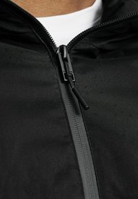 Svart zip-up jacka med en slät textur, som har en hög krage, en central dragkedja och dragkedjejustering vid huvan.