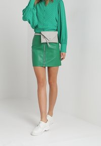 Blouse verte à motifs avec des manches ballons, associée à une jupe en cuir verte avec une fermeture éclair à l'avant. Accessoirisée d'un sac banane gris clair.