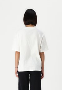 White cotton t-shirt with short sleeves and a round neckline, showing a relaxed fit from the back, paired with black pants.
