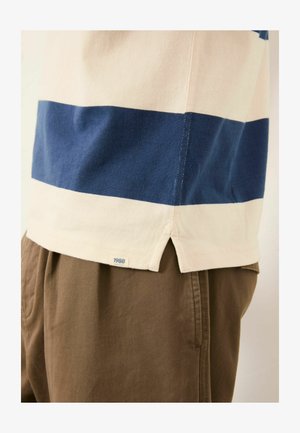 Gestreiftes Kurzarmhemd aus Baumwolle in Creme und Marineblau, mit seitlichem Schlitz und Etikett "1988", kombiniert mit braunen Stoffhosen.