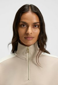 Pullover beige con zip e colletto alto, con finiture in oro e dettagli leggeri sulle spalle. Texture morbida con una finitura liscia.