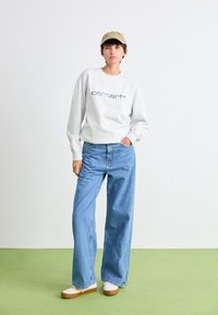 Light gray sweatshirt with printed "carhartt" logo, blue wide-leg jeans, beige cap, and white shoes with a tan sole on green floor.