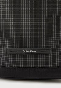 Bolso negro con diseño de cuadrícula, hecho de material sintético. Presenta una textura suave y una etiqueta con el logo de Calvin Klein.