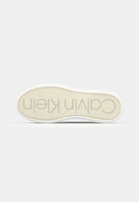 Semelle d'une basket blanche Calvin Klein montrant un motif de crampons avec le logo CK répété et le texte "Calvin Klein" au bas.