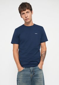 Marineblaue Baumwoll-T-Shirt mit kurzen Ärmeln, Rundhalsausschnitt und einem kleinen Logo auf der linken Brust. Getragen mit hellblauen Jeans.
