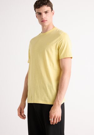 Yellow short-sleeve T-shirt made of lightweight fabric with a round neckline and a subtle logo. Worn with black pants.