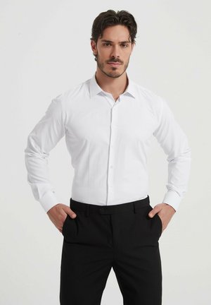 Chemise blanche à manches longues avec un devant à boutons, un col pointu et des poignets noirs contrastants, associée à un pantalon noir ajusté.