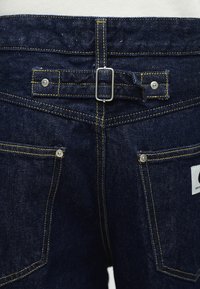 Πίσω μέρος σκούρων μπλε τζιν με αντίθετη κίτρινη ραφή, ασημί ριβέτ, δύο τετράγωνες τσέπες και μια κεντρική ρυθμιζόμενη λουρίδα με πόρπη.