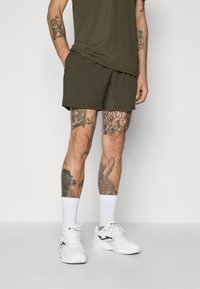 Olijfgroene sportshorts met een gladde textuur, gecombineerd met witte enkelsokken en witte sneakers. Opvallende tatoeages op zichtbare benen.