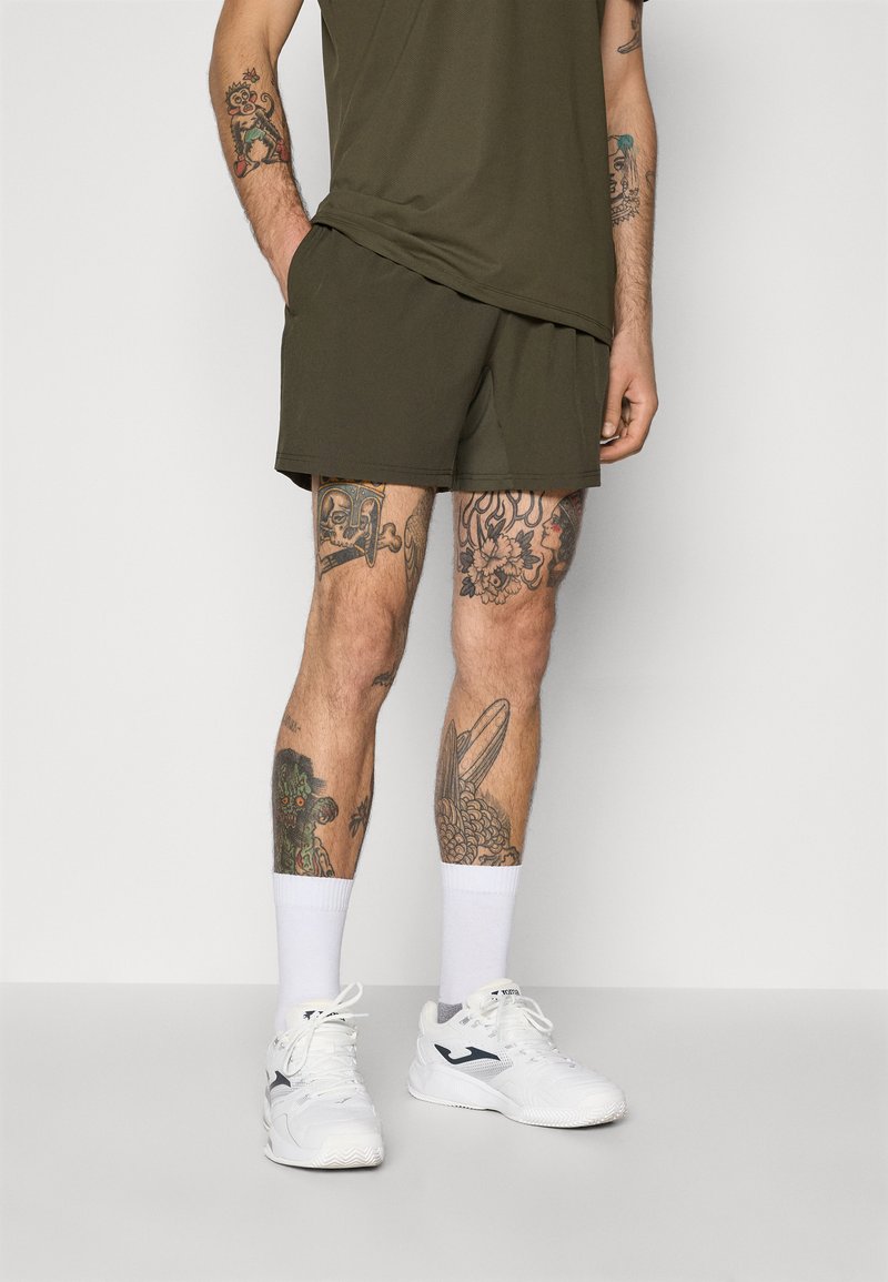 Olijfgroene sportshorts met een gladde textuur, gecombineerd met witte enkelsokken en witte sneakers. Opvallende tatoeages op zichtbare benen.