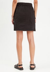 Jupe noire courte avec une taille élastique, présentant des coutures plates et une texture lisse, associée à des sandales noires.