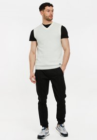 Helles graues, ärmelloses Oberteil mit V-Ausschnitt, ergänzt durch ein eng anliegendes schwarzes T-Shirt darunter und schwarze, schmal zulaufende Hosen, kombiniert mit grau-blauen Sneakers.