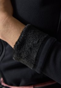 Crna majica dugih rukava s detaljima od teksturirane krzno na manžetama. Glatko tijelo od tkanine i mekano crno krzno na manžetama stvaraju kontrast u teksturi.