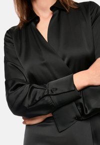 Zwarte satijnen wikkelshirt met een kraag, lange mouwen en knoopaccenten bij de manchetten. Soepele textuur en strak design.
