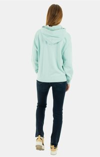 Felpa verde menta con cappuccio e vestibilità rilassata, dotata di tasca a marsupio e polsini a costine, abbinata a jeans blu scuro e sneakers di colore chiaro.