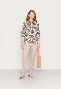 Blusa patterned em preto e branco com mangas até os três quartos, calças beige ajustadas e uma bolsa de palha. As sandálias têm um design decorativo.