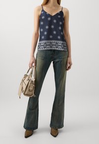 Marinblå topp med vita blommönster, utsvängda mörk denimjeans och en beige handväska med dragkedjor och en liten etikett, som hålls med en hand.