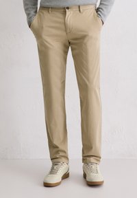 Beige cotton trousers with a straight cut, front pockets, and a zipper fly, paired with light-colored sneakers and a grey sweater.