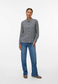 Kvinde iført en langærmet sort og hvid vertikalstribet skjorte, blå jeans og brune fritidssko, stående foran en hvid baggrund.