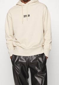 Beżowa bluza z kapturem z kieszenią kangurką z przodu, sznurkiem przy kapturze i czarnym haftem "DRYARN". Połączona z czarnymi, wysokim stanem, spodnie z imitacji skóry.